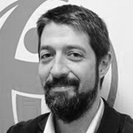 Et svart-hvitt portrett av en mann med bart og skjegg, iført dress og slips, som smiler.