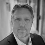 Et svart-hvitt portrett av en mann med kort hår, iført dress og skjorte, står innesluttet i en lys bakgrunn.