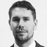 En svart-hvitt portrettfotografi av en mann med mørkt, kort hår og skjegg, iført en dress og skjorte.