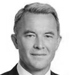 En mann iført mørk dress og hvit skjorte, smilende.