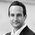 Dette er et svart-hvitt portrett av en mann i dress, smilende, med kort mørkt hår og lys hud.