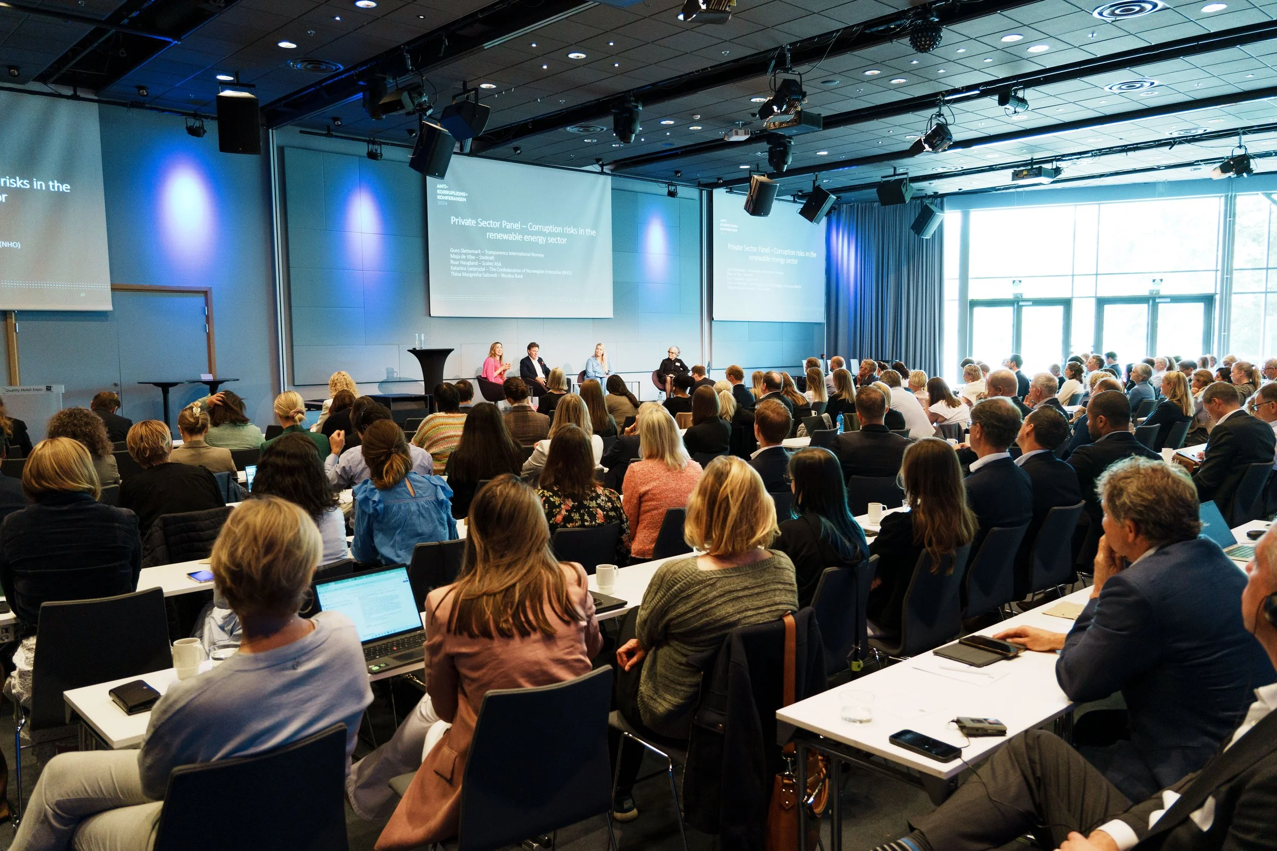 Et konferanserom med mange mennesker som lytter til en paneldebatt på scenen. Det er fire personer som snakker, og flere visuelle skjermer viser presentasjonsmateriale.