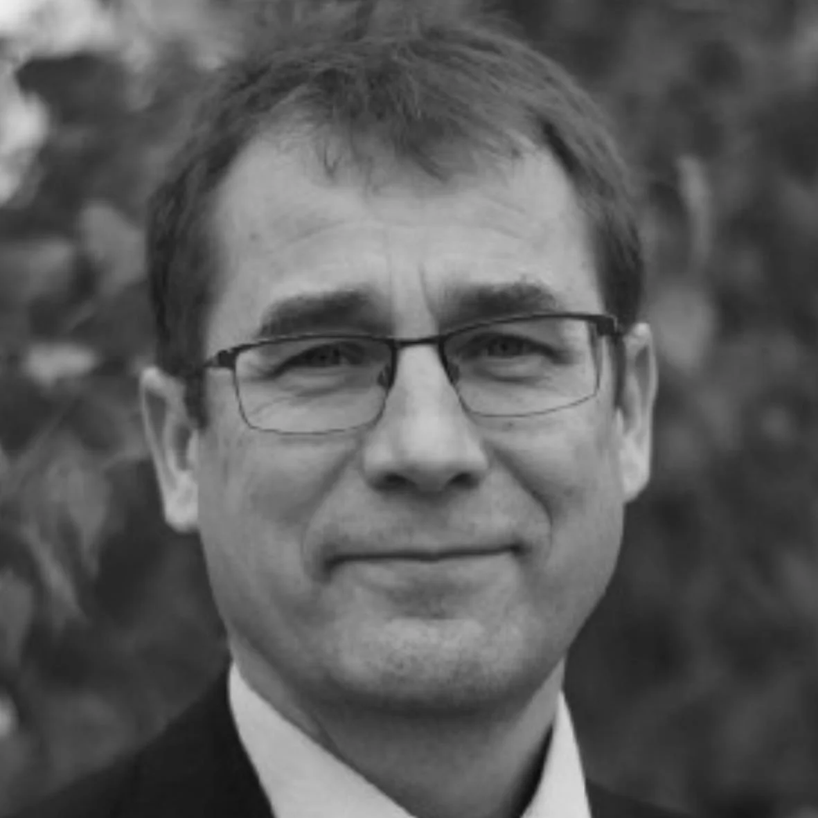 Portrett av en mann med briller, kort brunt hår, iført sort dress og hvit skjorte, uten farge, med bakgrunn av blurry trær.