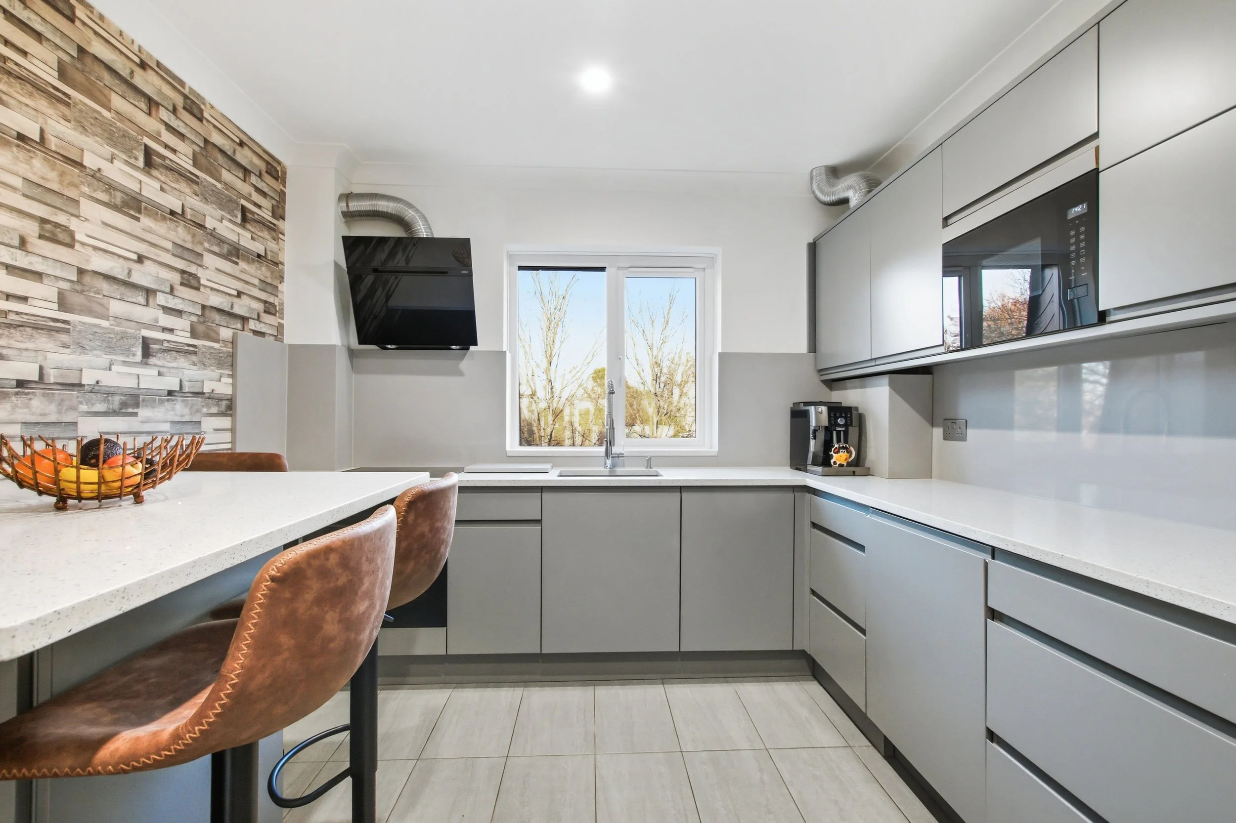 Modern kitchen with grey cabinets, white countertops, and a window. A wall with textured wood paneling features a black vent hood. There are two brown bar stools at a white counter, a basket of fruit, a coffee machine on the counter, and a microwave above. The flooring is light-colored tile.