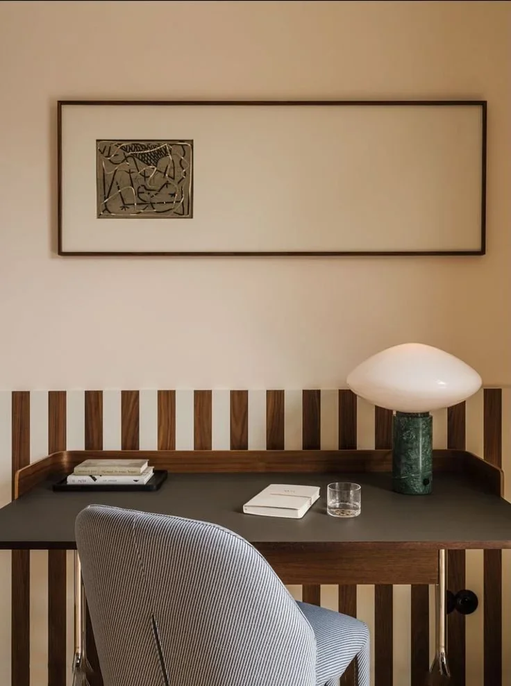 A home office workspace with a wooden desk, striped gray chair, modern table lamp, books, and a glass of water, with a framed abstract artwork on the wall.