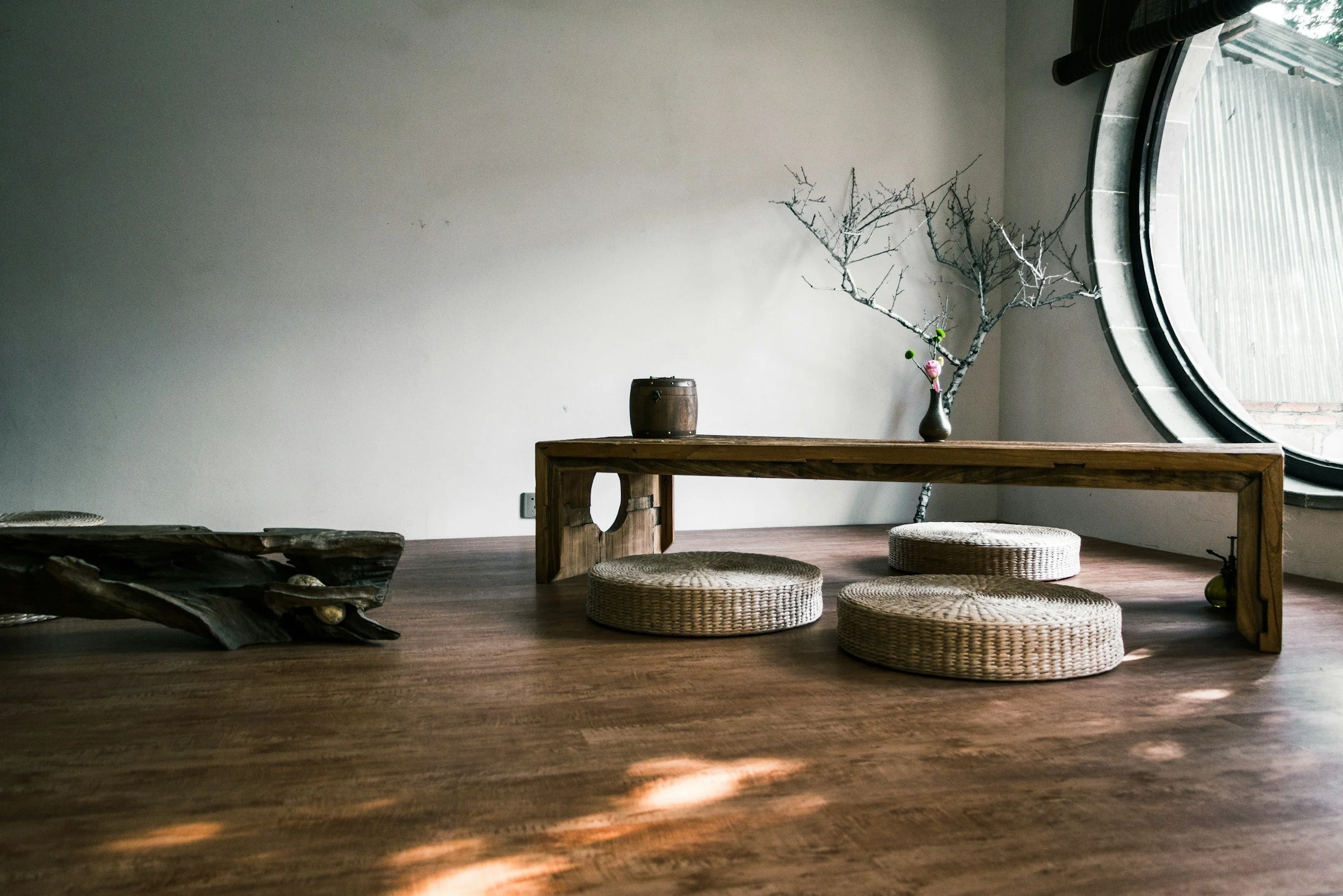 Minimalist indoor space with a low wooden table, round woven cushions, and a decorative leafless branch in a vase, near a large circular window.