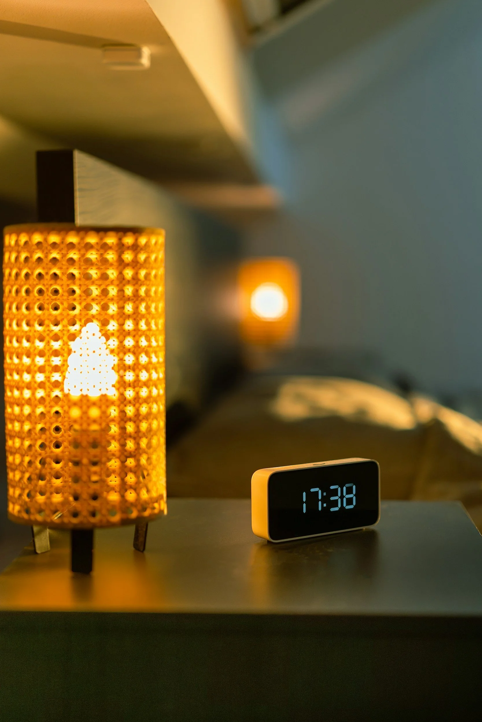 A digital clock on a bedside table shows the time 17:38, next to a rattan-style lamp with a warm glow in a bedroom setting.