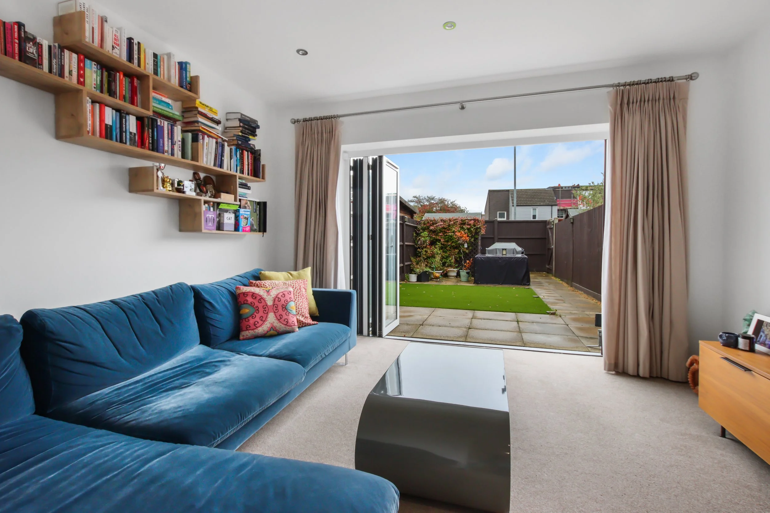 Modern living room with blue sectional sofa, colorful cushions, wall-mounted bookshelves filled with books, large glass doors opening to an outdoor patio with a garden, and a glass coffee table.
