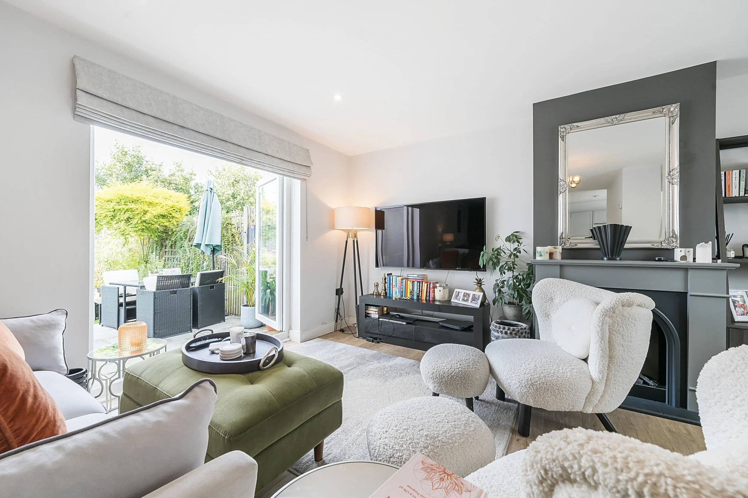 Modern living room with large TV, cozy seating, and patio access. Features a gray accent wall, mirror, decorative fireplace, bookshelf, and floor lamp. Sliding glass doors open to an outdoor area with patio furniture and greenery.