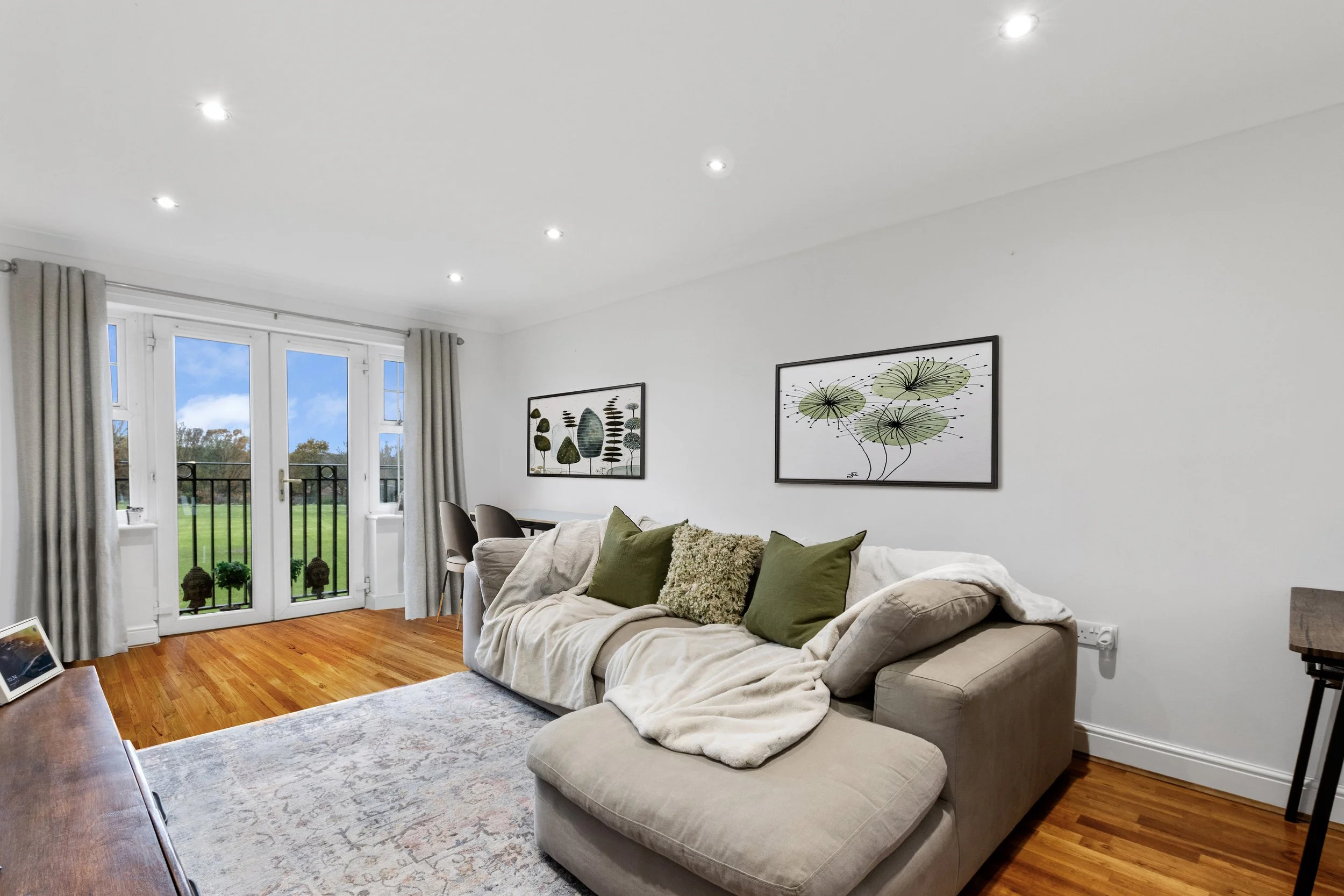 Modern living room with beige sofa and green cushions, wooden floor, and white walls featuring framed botanical artwork. There is a rug on the floor, a small dining table with chairs in the corner, and large French doors with curtains opening to an outdoor area.