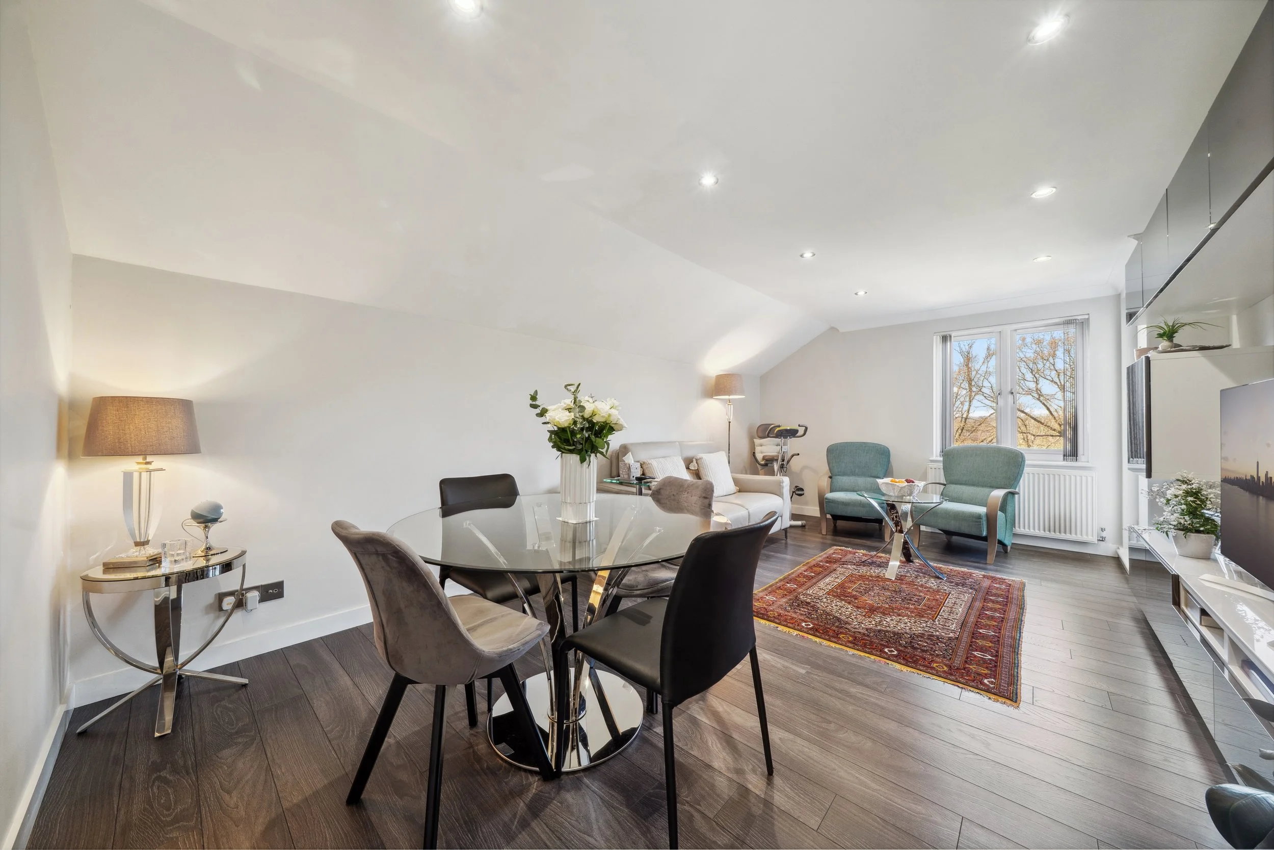 Modern living room with glass dining table, four chairs, sofa, two armchairs, exercise bike, and a flat-screen TV. A window overlooks a tree-filled landscape. The room is decorated with a decorative rug, side tables, lamps, and vases with flowers.
