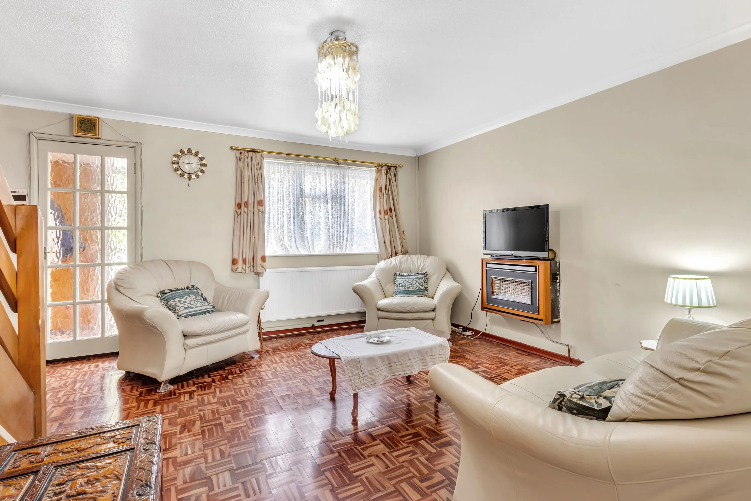Living room with cream leather sofas, wooden parquet floor, window with floral curtains, wall-mounted TV and electric fireplace, glass door, and a ceiling light fixture.