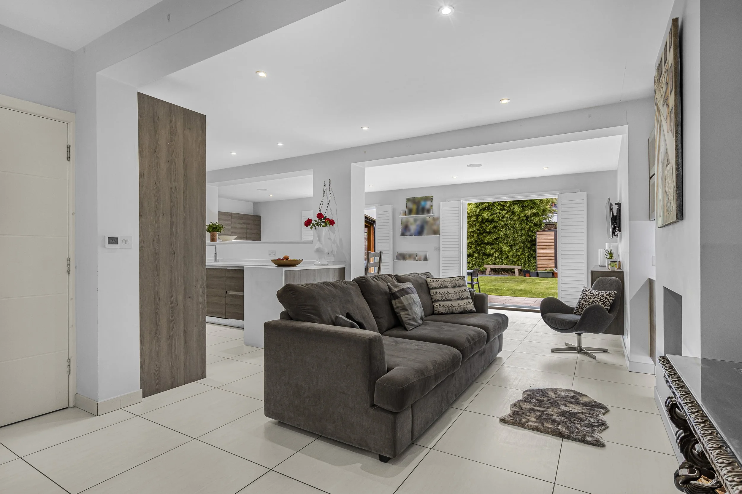 Modern living room with brown sofa, black swivel chair, olive-green wall, and open patio door showing a backyard garden.