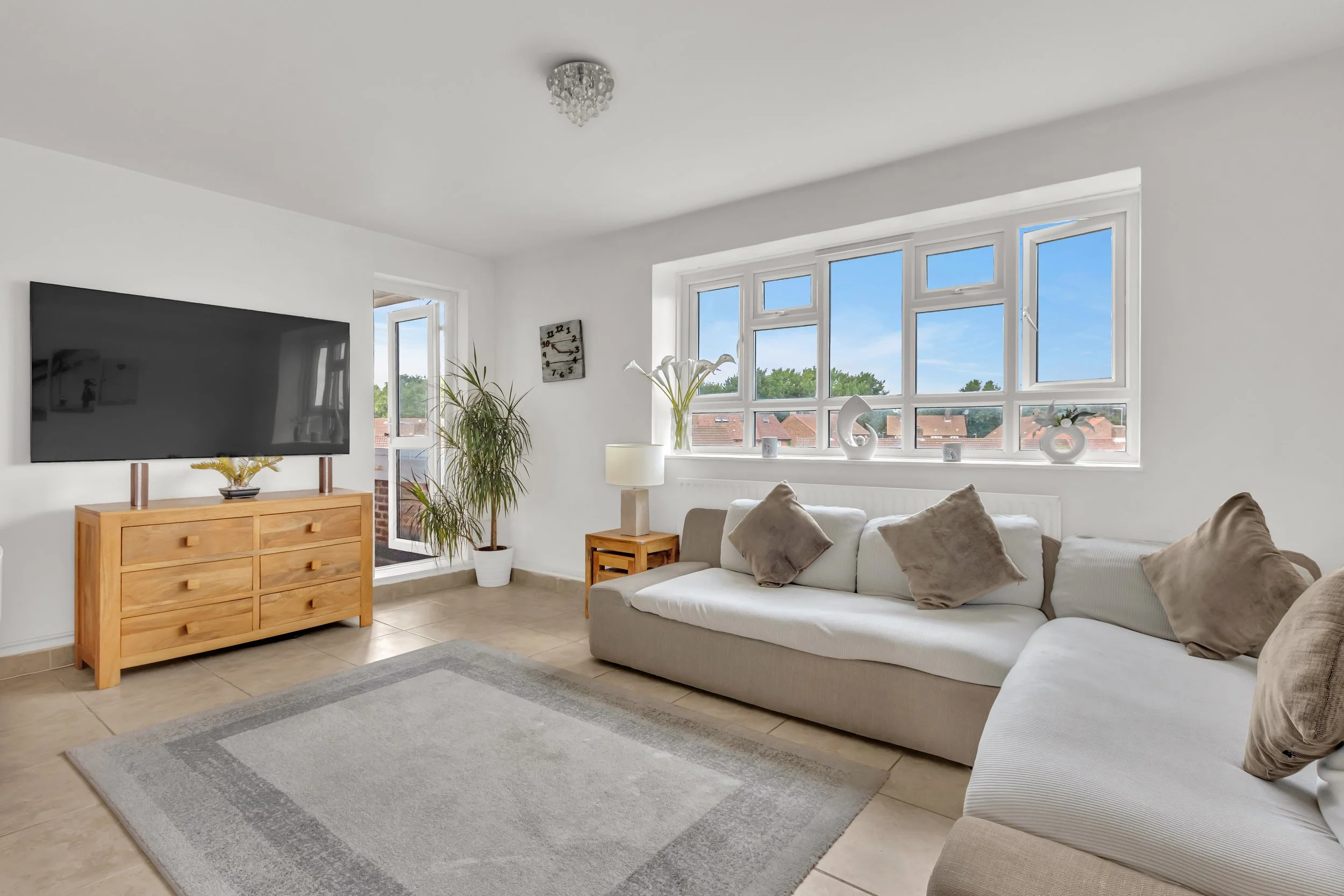 Bright living room with white walls, beige sectional sofa with brown and beige pillows, large window with outside view of houses and trees, wooden TV stand with a flat-screen TV, potted plant, small side table with a lamp, decorative items on windowsill, today’s date card, light-colored tile floor and area rug.