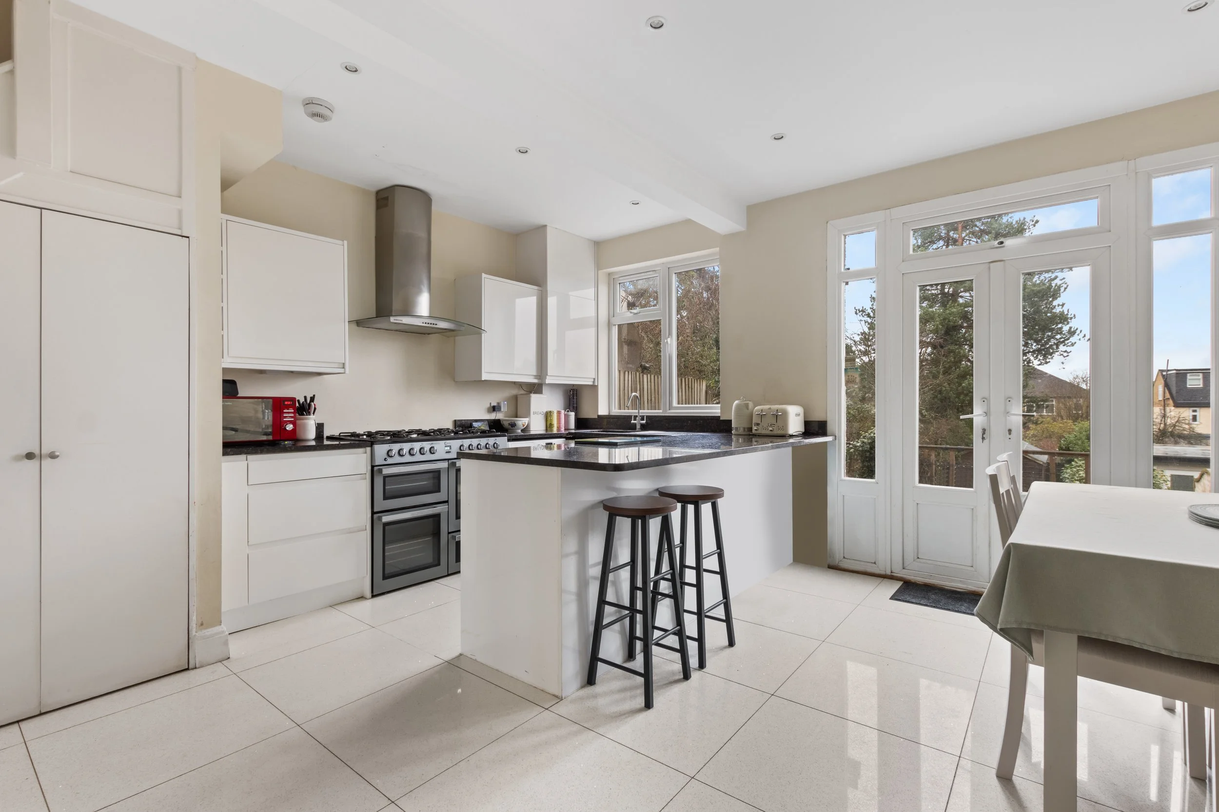 Modern kitchen with white cabinets, black countertops, a gas stove, a double oven, a stainless steel range hood, and large windows letting in natural light, with a door leading outside.