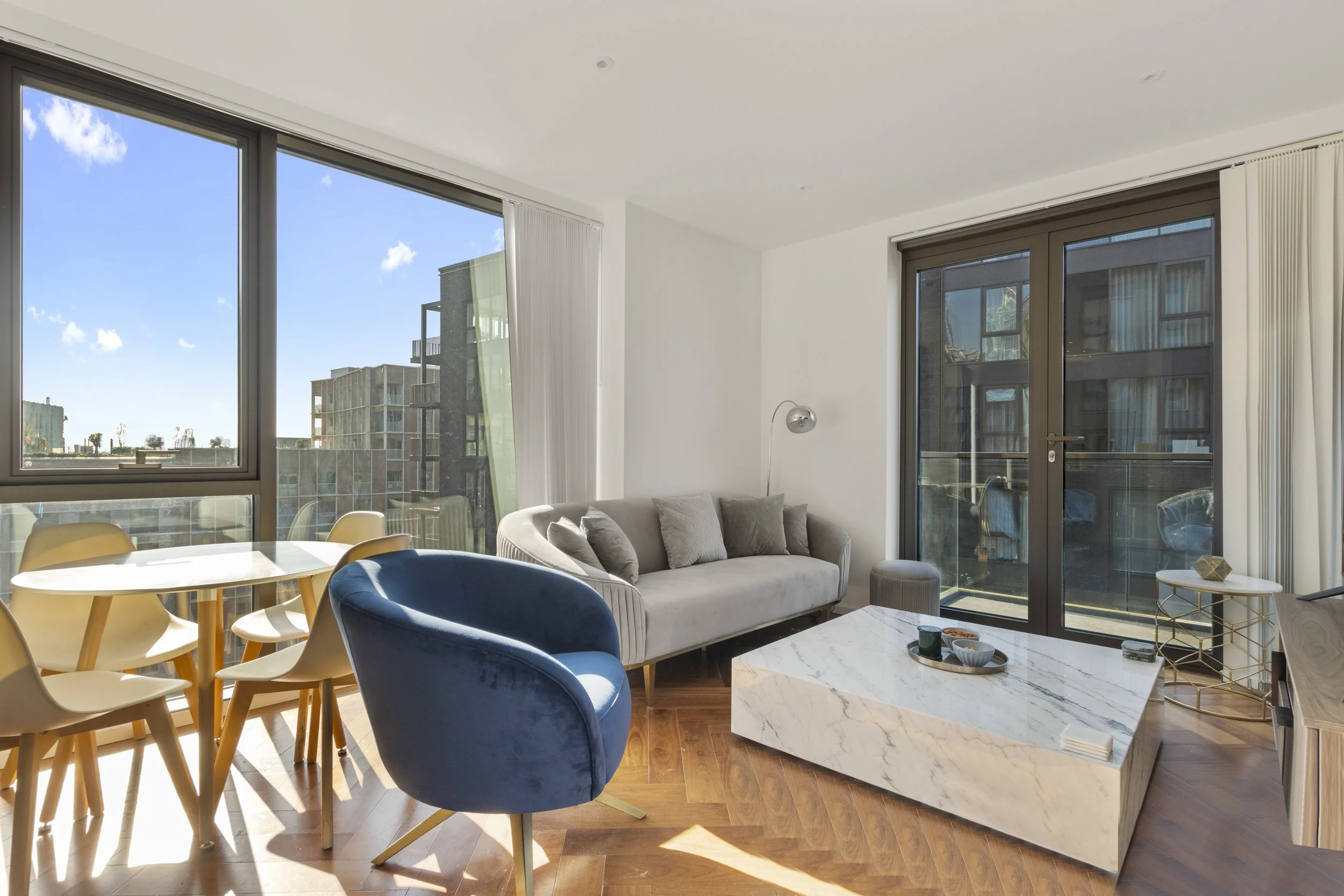 Modern living room with large windows, sofa, blue velvet accent chair, marble coffee table, dining table with yellow chairs, and city view outside.