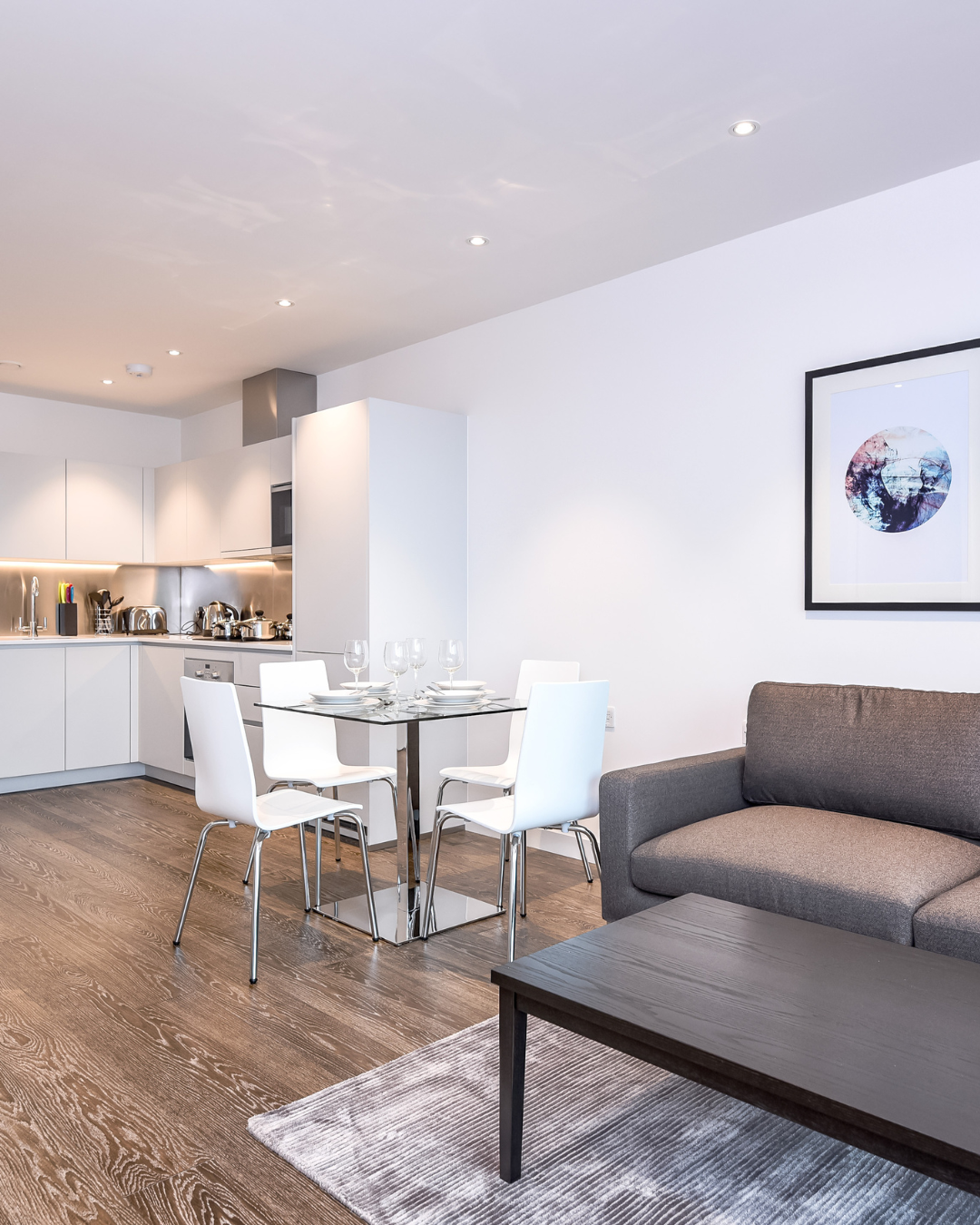 Modern open-plan living and dining area with a kitchen, featuring a gray sofa, a black coffee table, a dining table with white chairs, and a framed picture on the wall. The kitchen has white cabinets and stainless steel appliances.