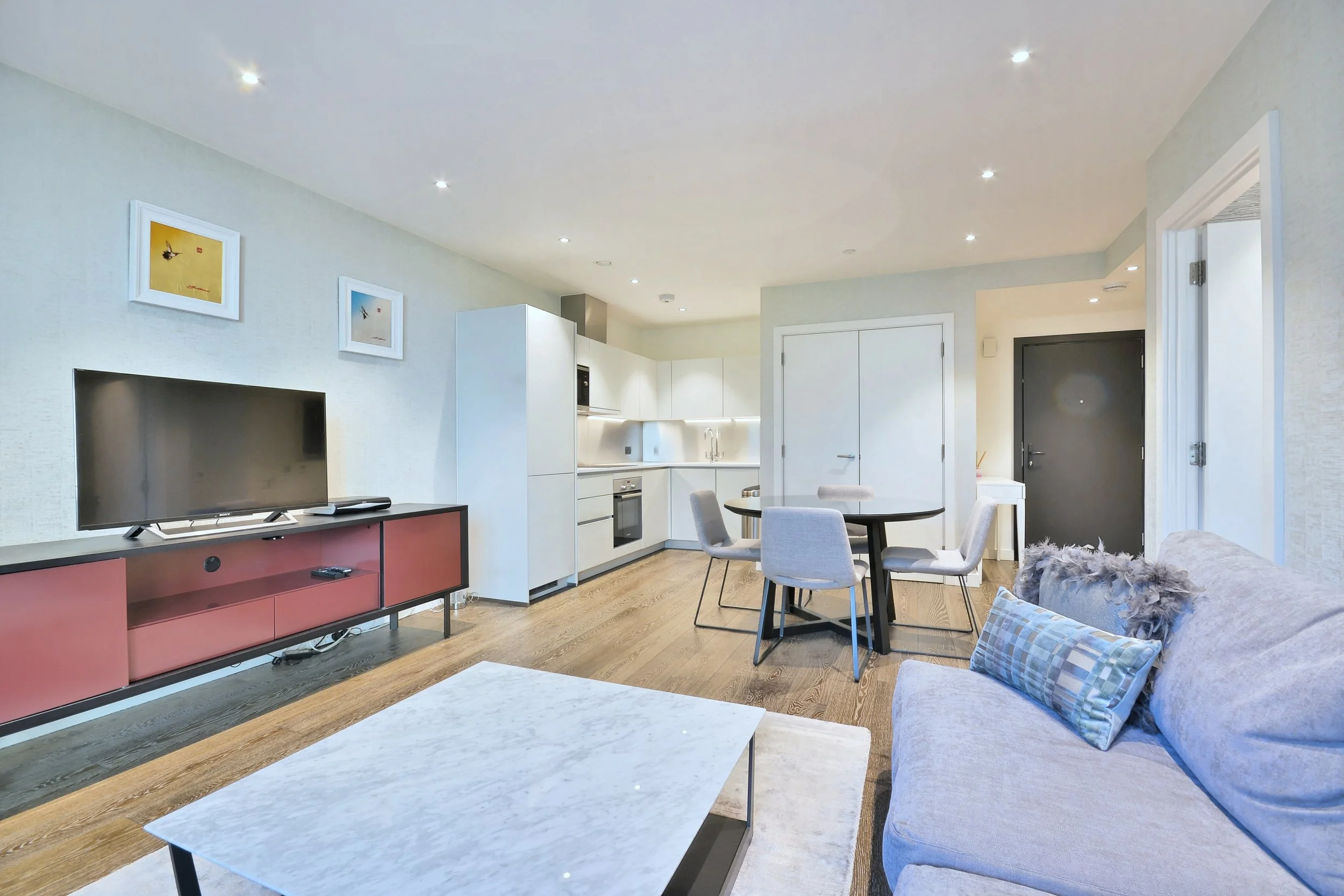 Modern living room with a TV, red media console, gray sofa, round dining table with gray chairs, minimalist kitchen, and light hardwood flooring.