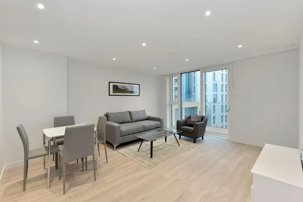Modern living room with gray sofa, glass coffee table, armchair, dining table, and chairs, large windows, and wooden floor.