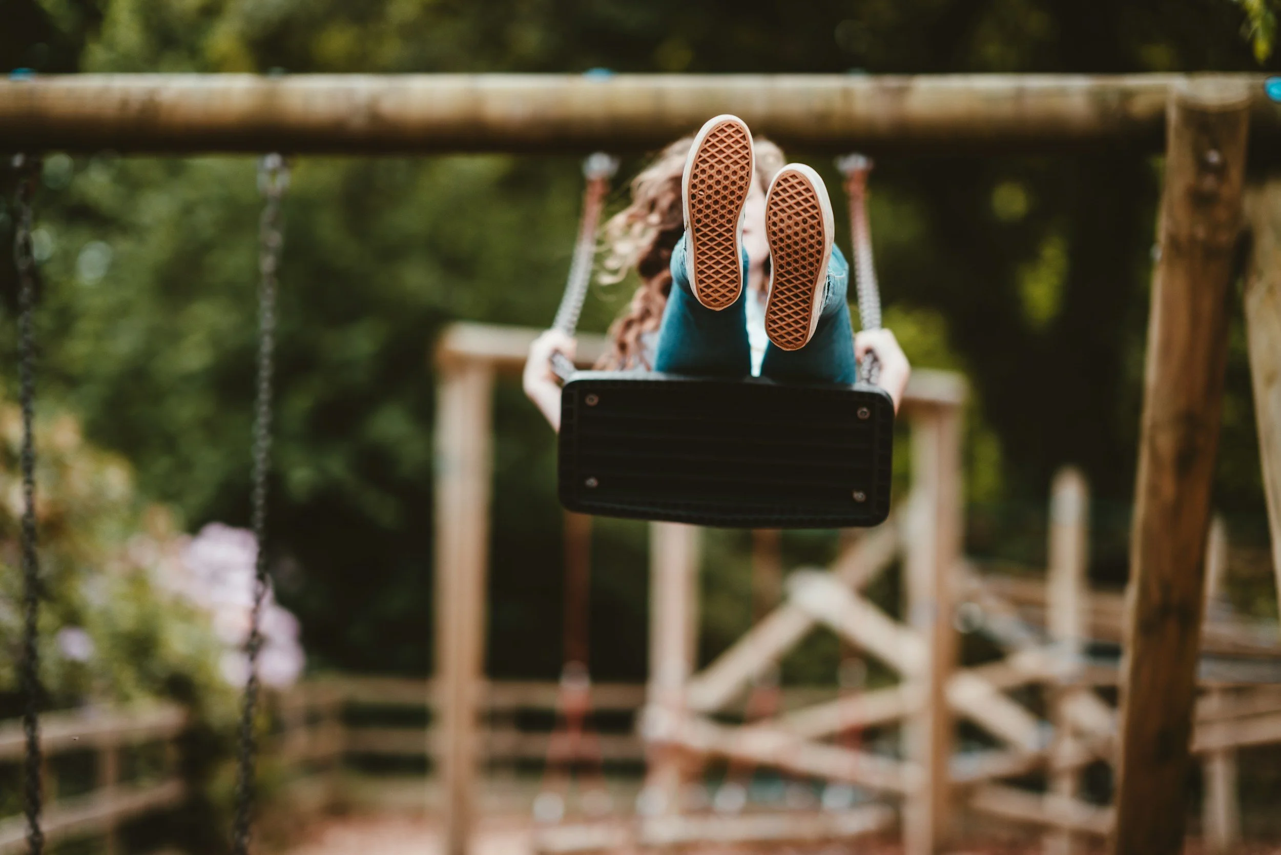 A person with long hair sitting on a swing at a playground surrounded by greenery, with the focus on the soles of their shoes.