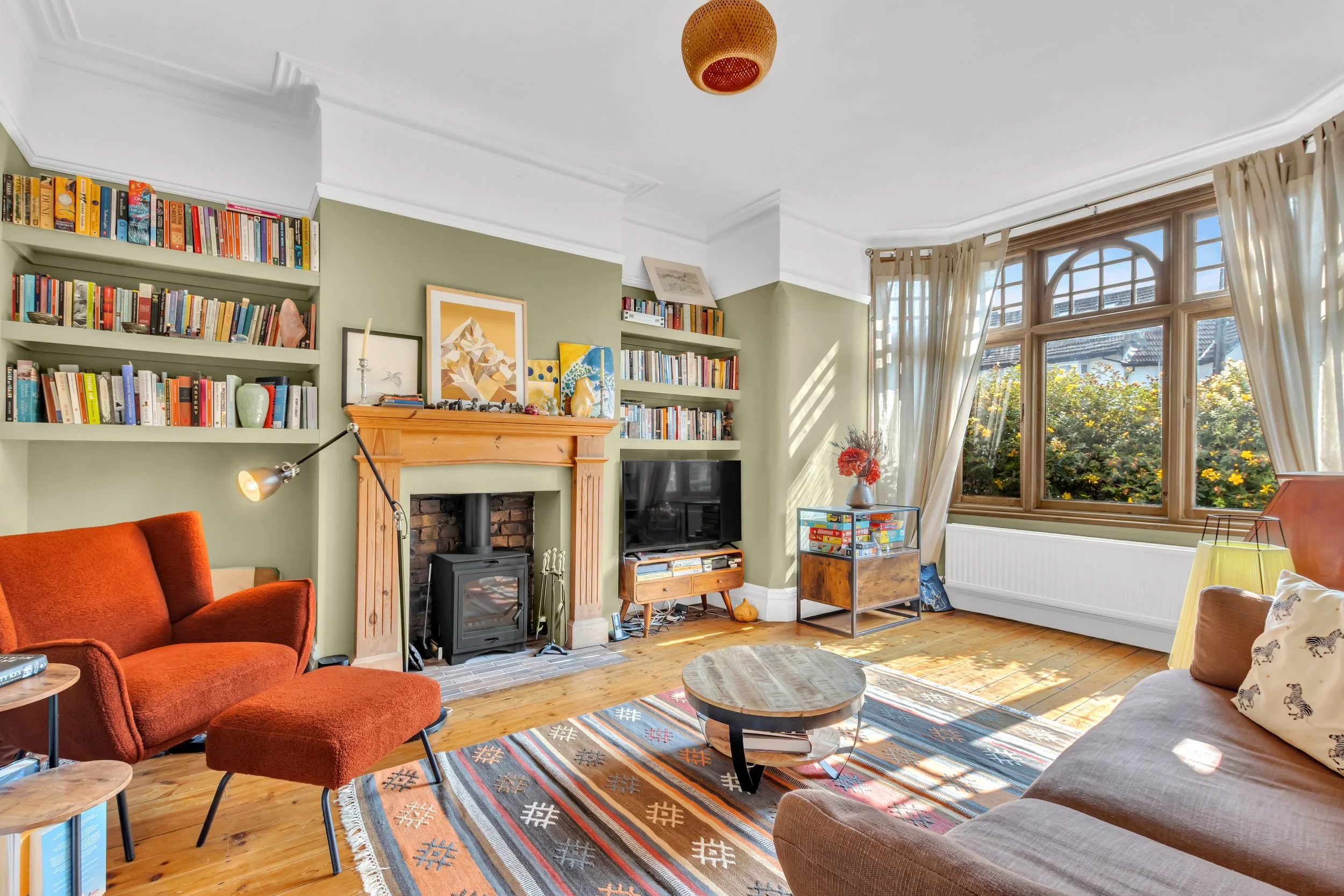 Living room with green walls, a fireplace, bookshelves, a flat-screen TV, a large window with curtains, a red sofa with a matching ottoman, a beige sofa, a round wooden coffee table, and a colorful rug.