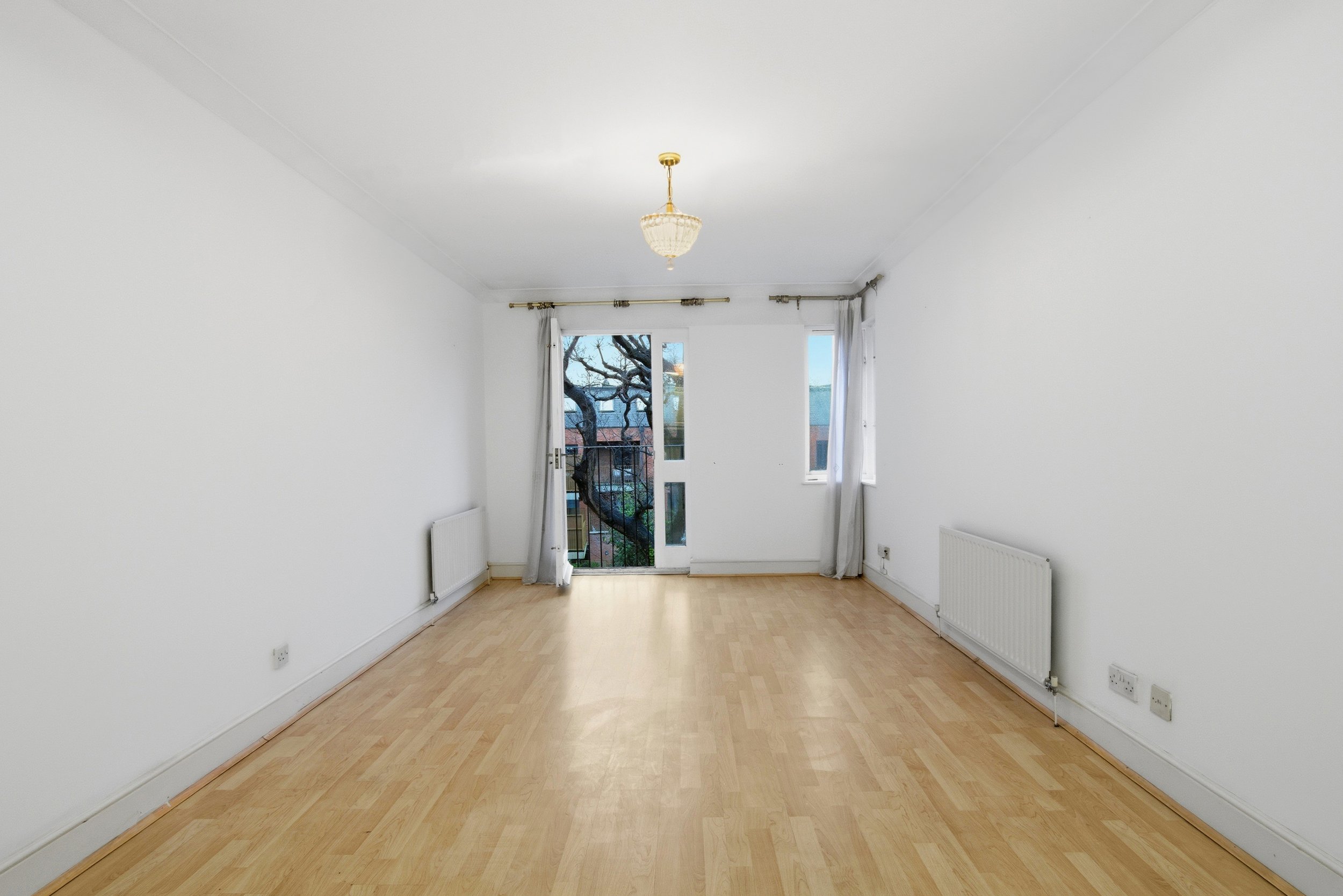 Empty living room with white walls, light wood flooring, a ceiling light,  and a large window with curtains, showing a tree outside.