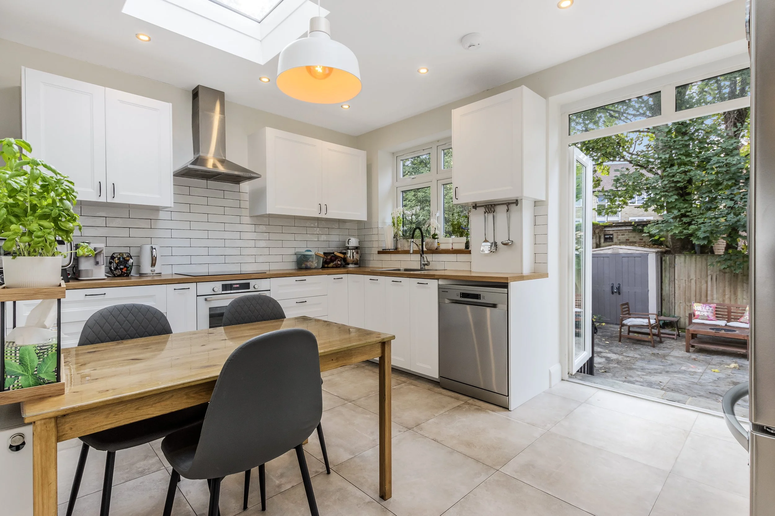 Bright modern white kitchen with subway tile backsplash, stainless steel appliances, wood dining table, pendant lighting, and open patio doors