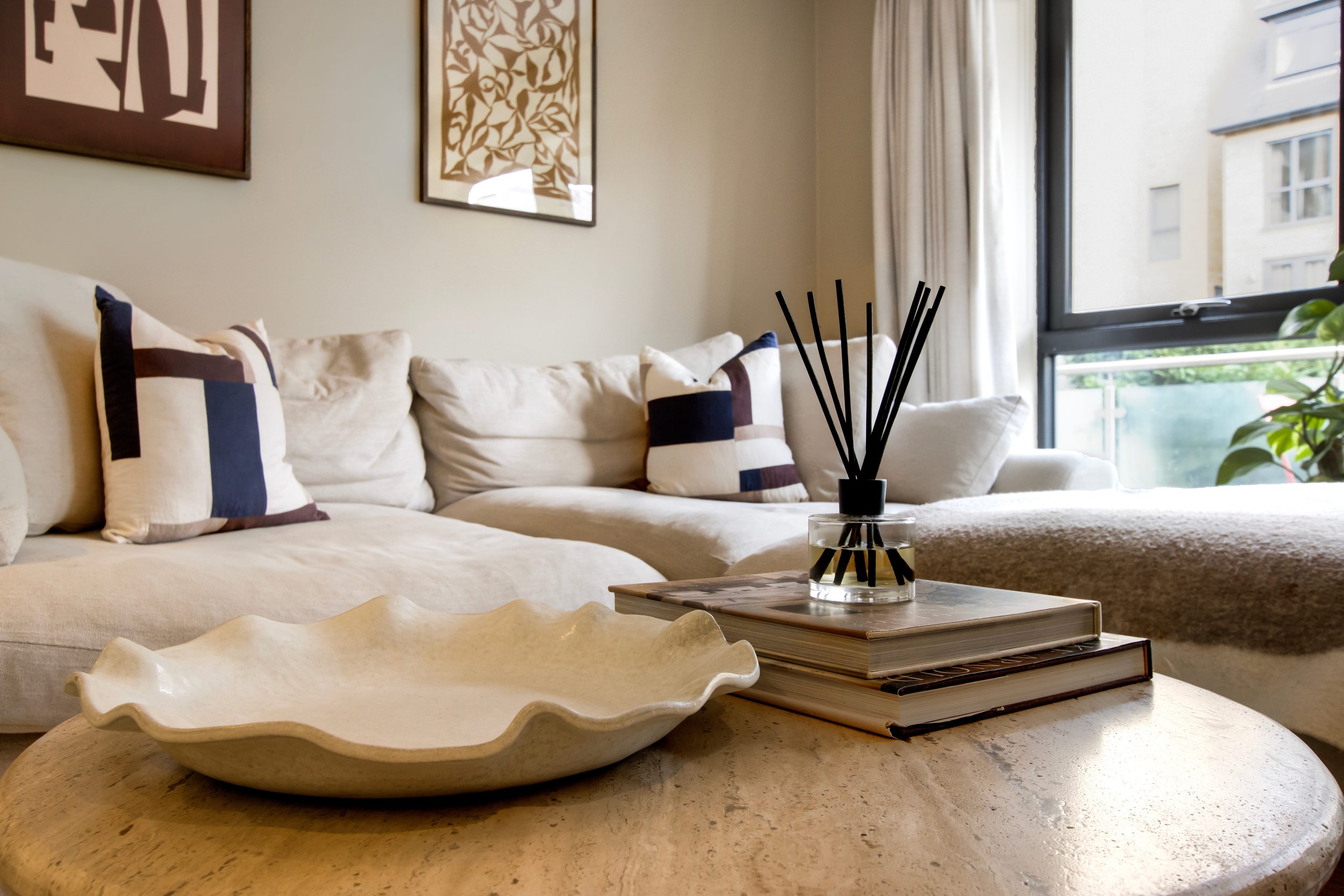 Bright modern living room with a cream sectional sofa, patterned pillows, reed diffuser, decorative books, sculptural bowl, wall art, and large window