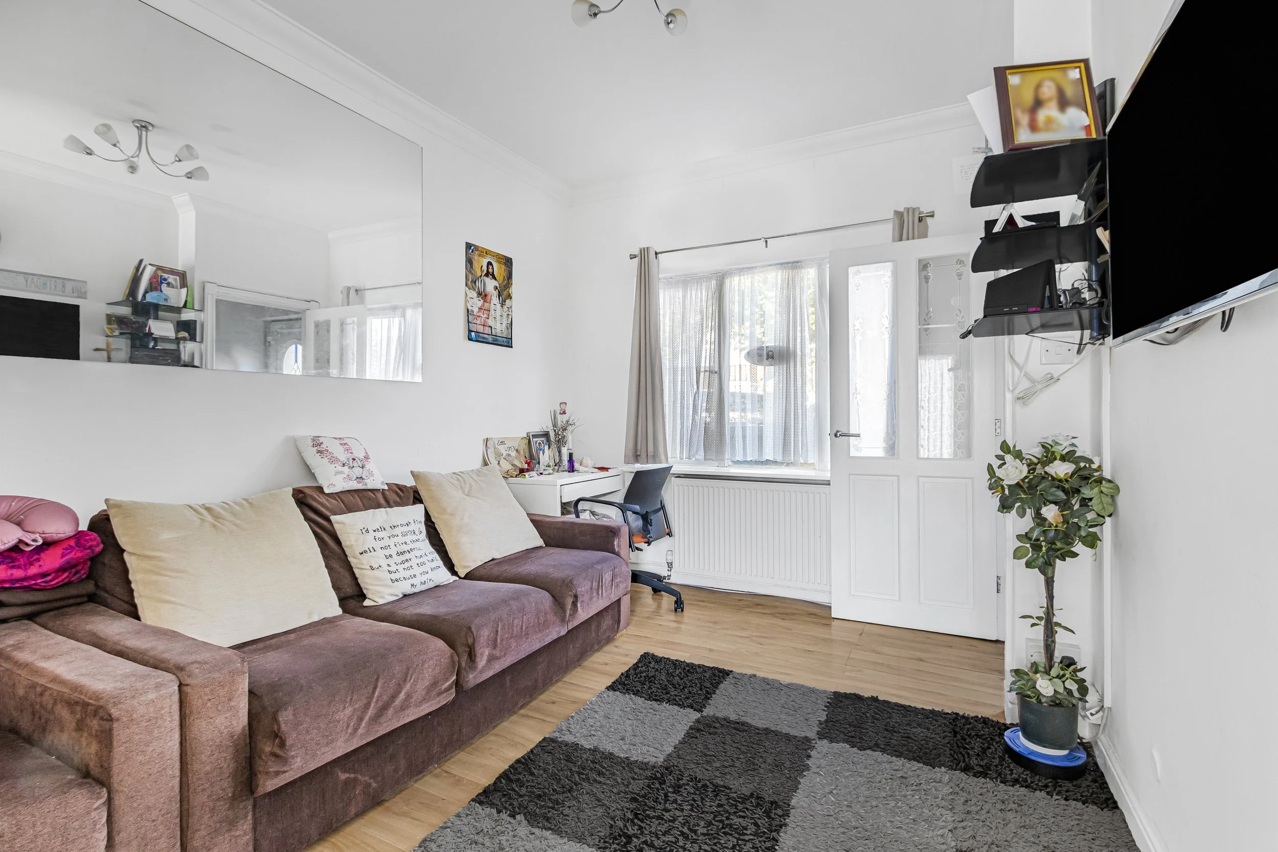 Living room with a brown sofa, pillows, a black rug, a white desk, a black office chair, a television mounted on the wall, a potted plant, and a window with curtains.
