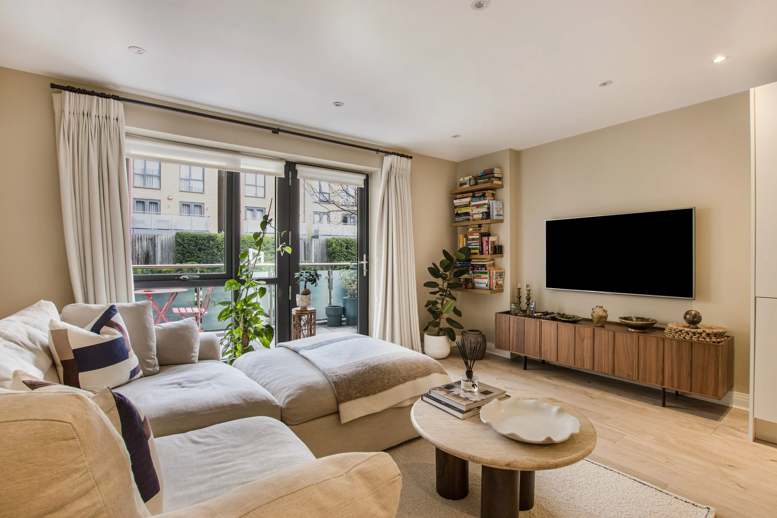 Living room with beige sofa, round wooden coffee table, wall-mounted flat-screen TV, bookshelves, indoor potted plants, and large windows with white curtains.