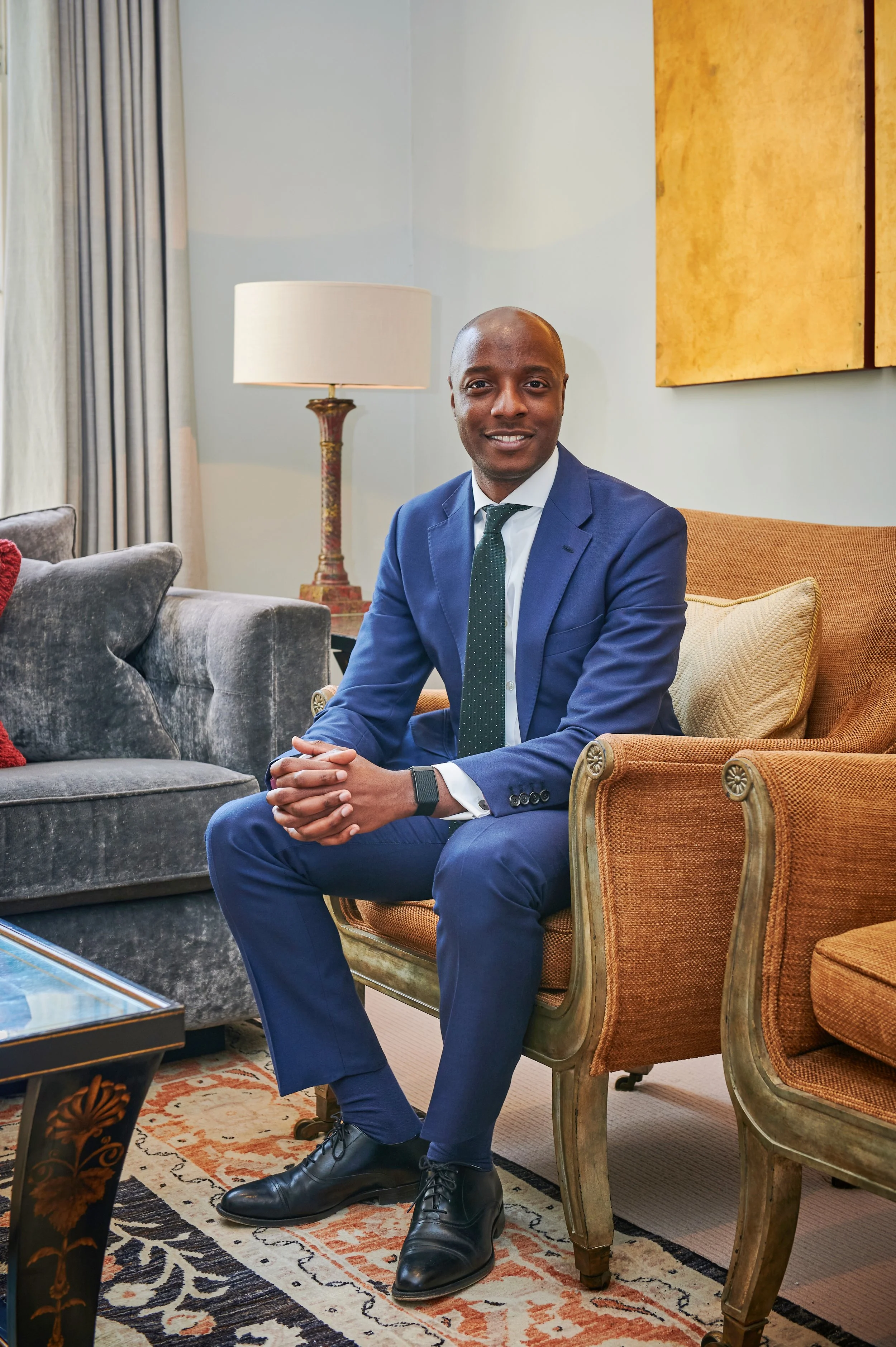 A man in a blue suit, white shirt, and dark green tie sitting on a vintage-style armchair in a well-decorated room with a gray sofa, a lamp, and artwork on the wall.