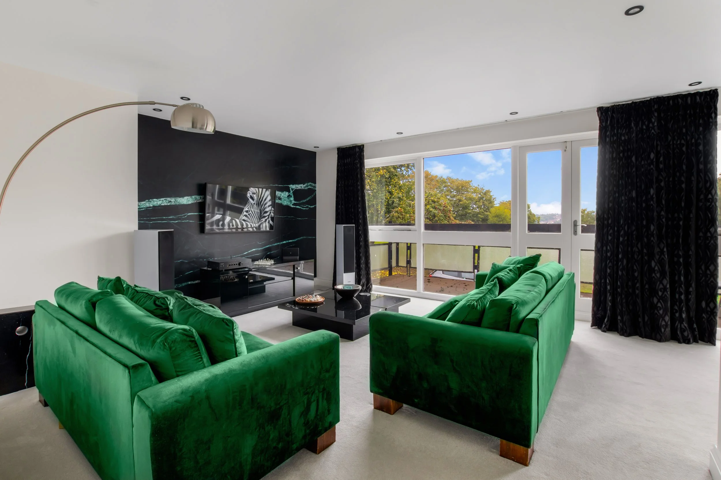 Living room with two green velvet sofas, a black coffee table, a black textured accent wall, a large windows with black curtains, and a balcony outside.