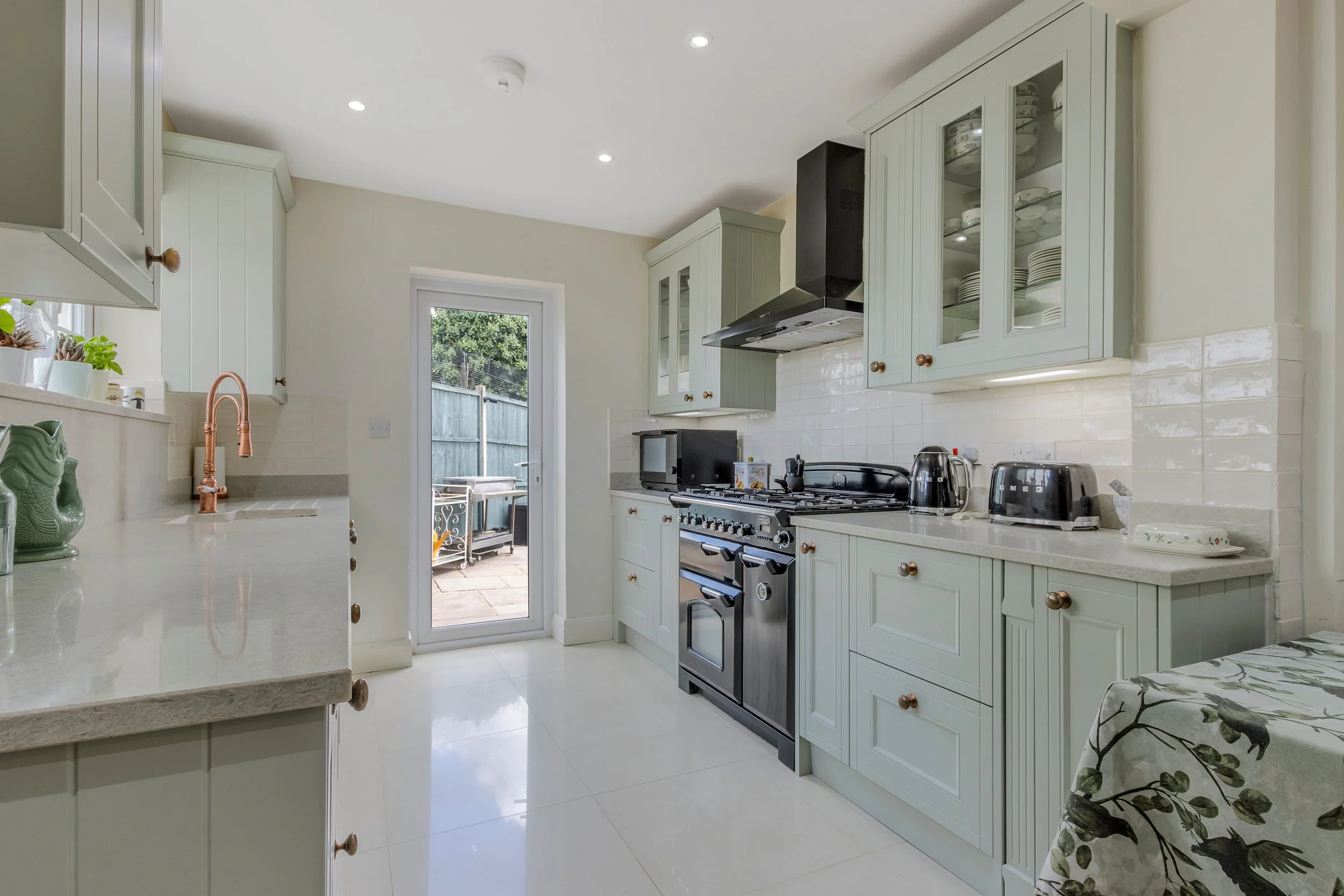 Bright traditional kitchen with pale green cabinets, glass-front dish cupboards, copper farmhouse faucet, gas range, black hood, and white countertops
