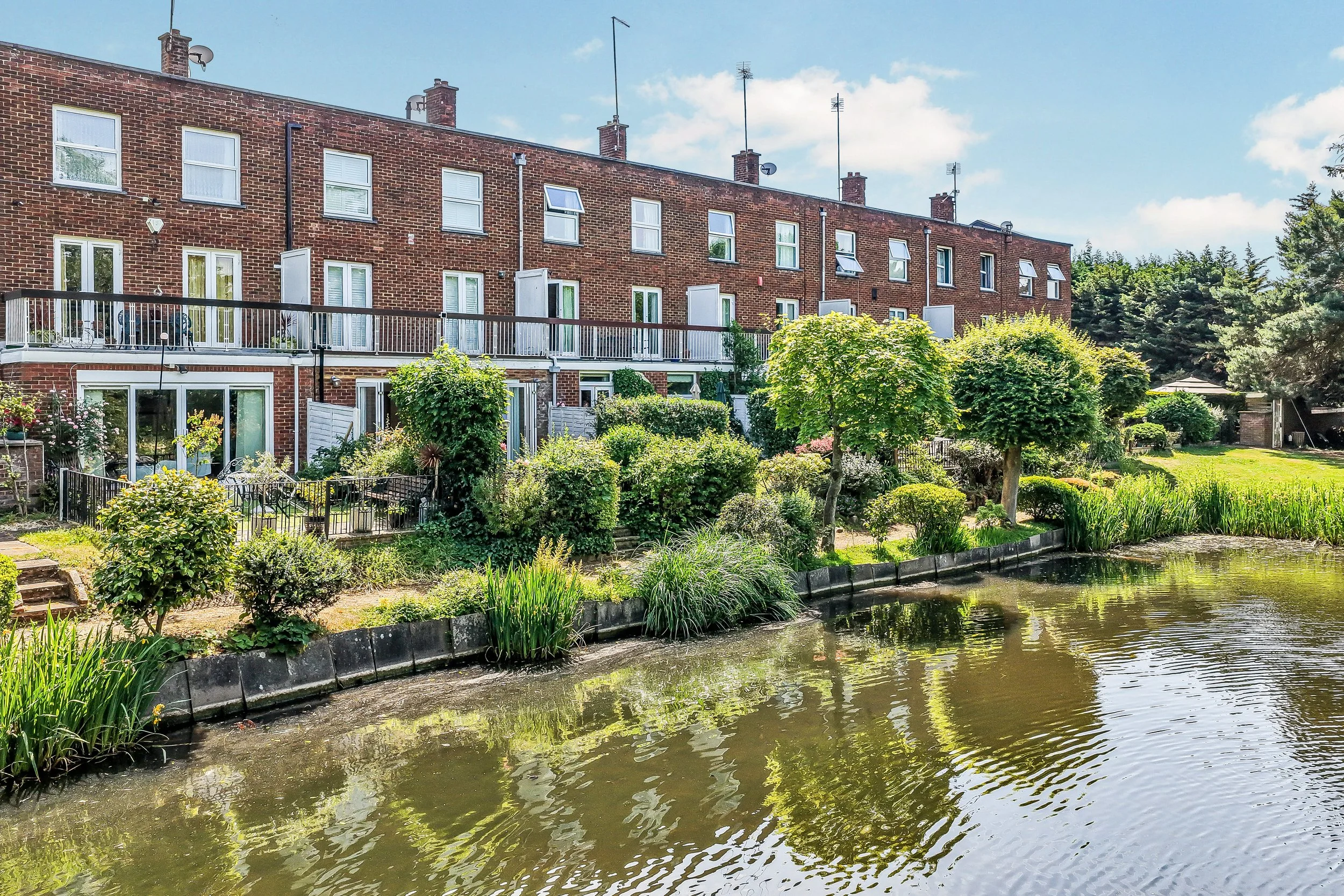 A brick apartment building with several small balconies, attached to a garden with various trees and flowering bushes, next to a pond with calm water reflecting the greenery underneath a partly cloudy sky.