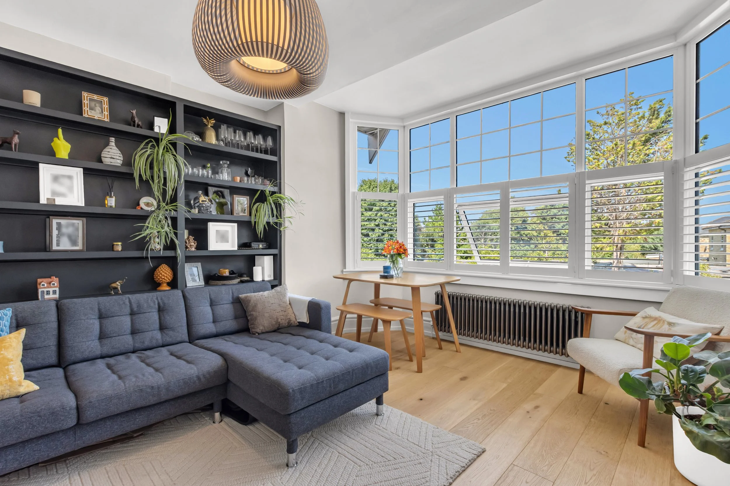 Bright living room with large bay window, gray sectional sofa, black built-in bookshelf with decorations, small dining table with flowers, cream armchair, and wooden flooring.