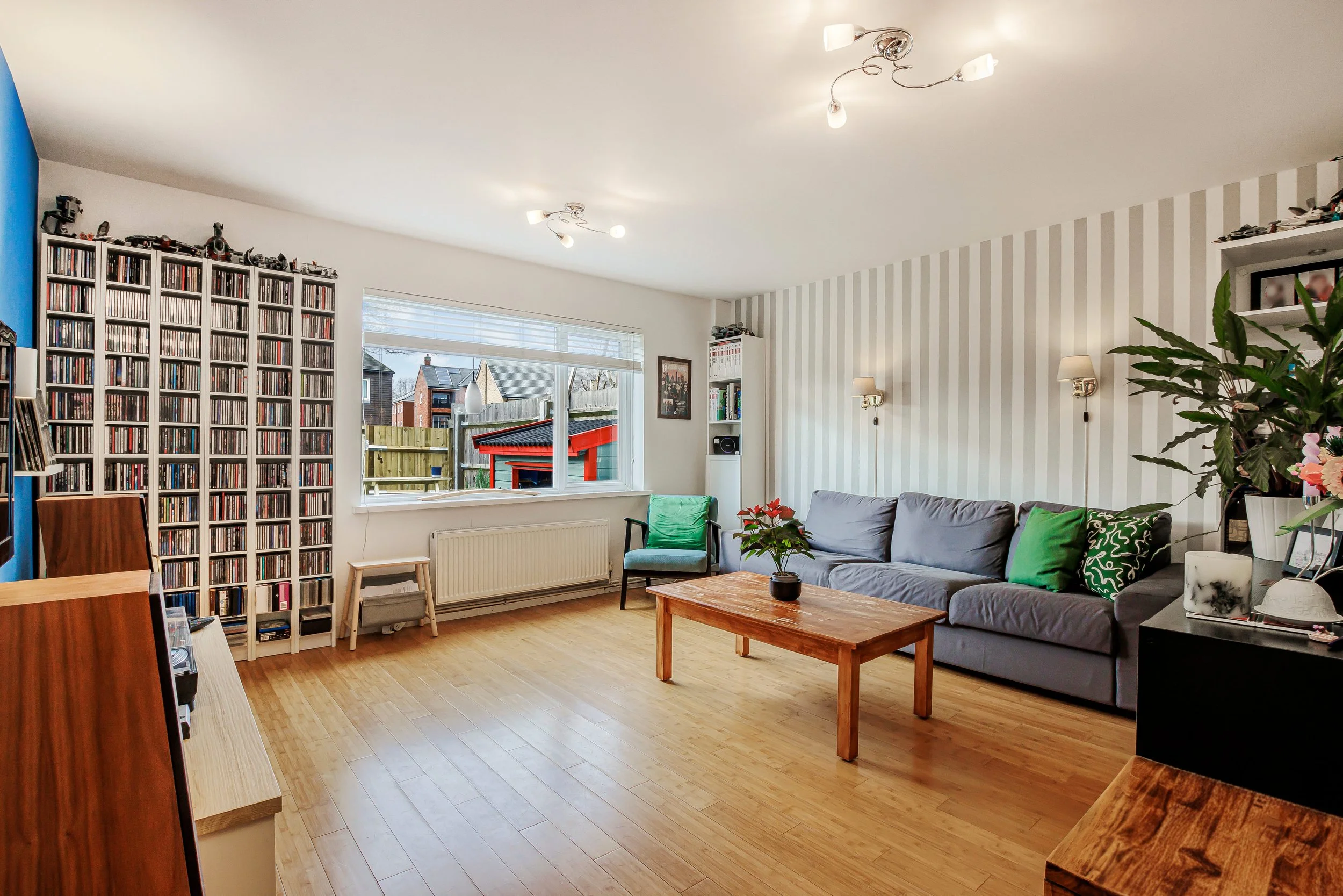 Living room with wooden floor, gray sofa, green cushions, wooden coffee table, armchair, large window, white and blue walls, bookshelf, plants, ceiling lights, wall lamps, and outdoor view.