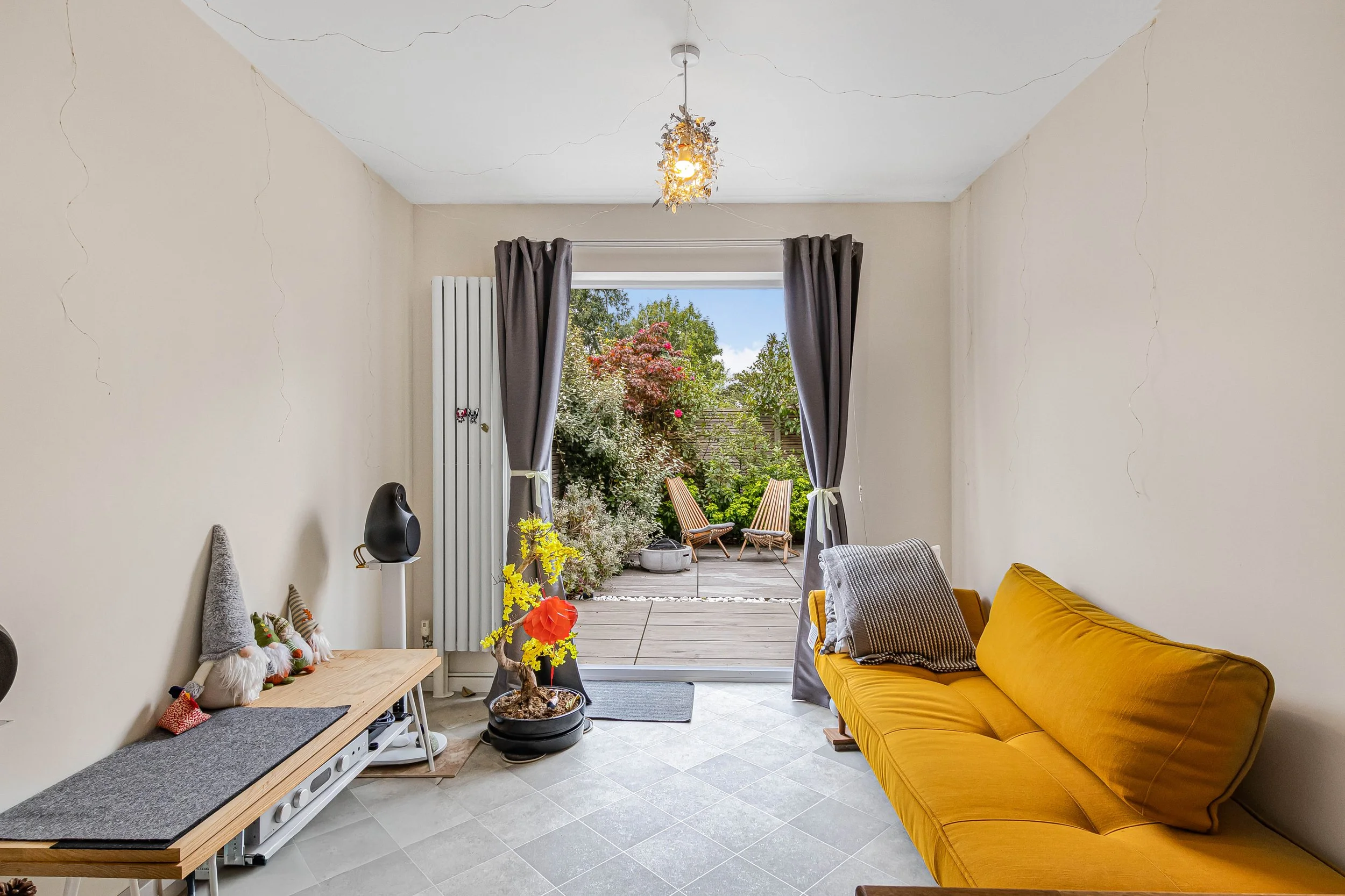 Living room with a yellow sofa, decorated with a pillow, leading to an outdoor patio with two wooden chairs and a garden with trees and pink flowers, seen through partially open gray curtains.