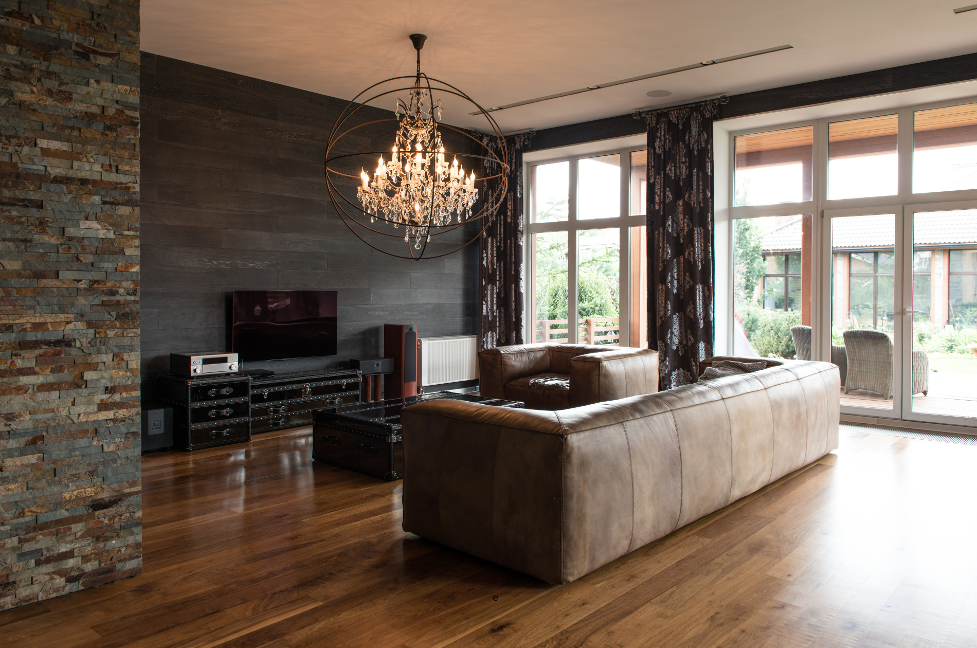 Modern living room with leather sofas, a chandelier, a flat-screen TV, and large windows with dark patterned curtains, overlooking a patio area.