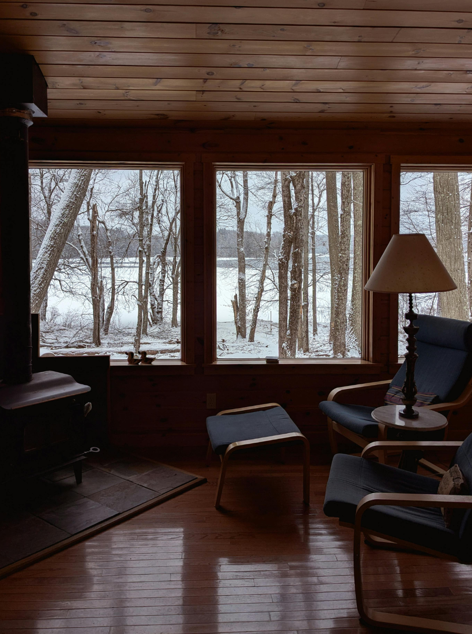 Interior view of a cozy room with wooden walls and ceiling, large windows showing a snowy landscape with trees outside, a rocking chair with a cushion, a small table with a lamp, a chair, and an ottoman.