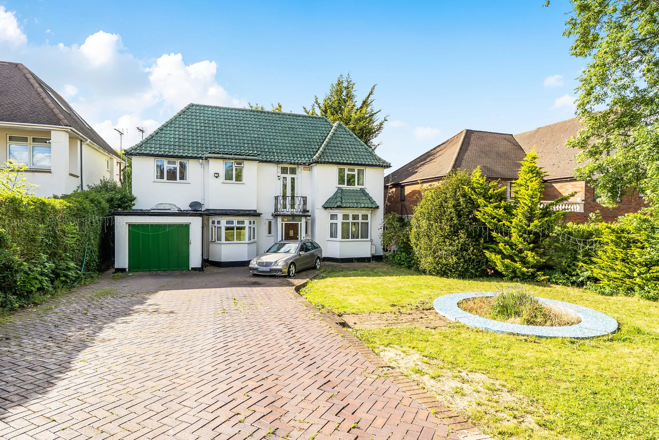 A white two-story house with a green tiled roof, green garage door, and a small front porch. A silver car is parked in the driveway. There are shrubs and trees surrounding the house, with a neatly paved brick driveway and a circular garden bed in the front yard. The sky is blue with some clouds.