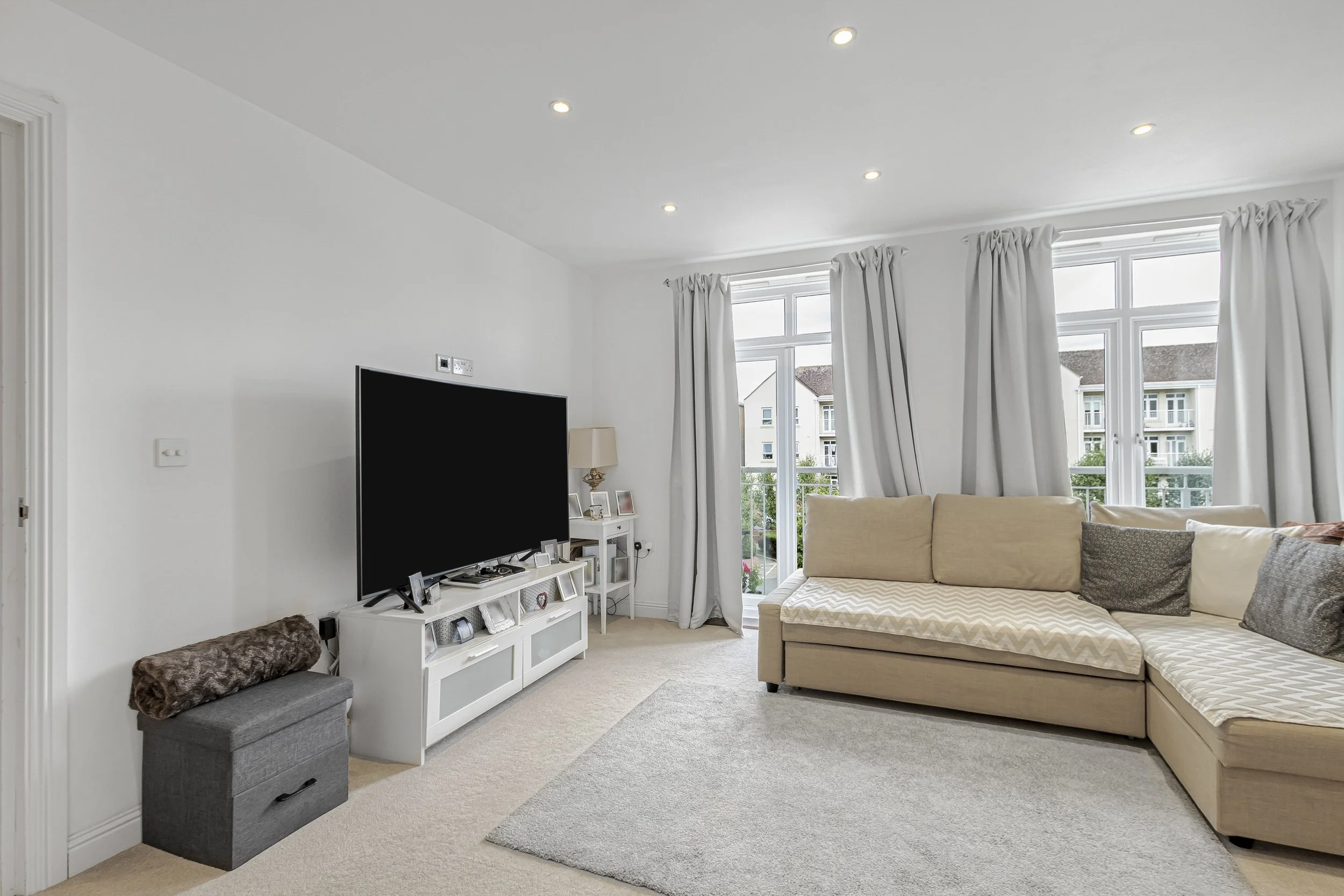 Bright living room with beige L-shaped sofa, large windows with white curtains, and a flat-screen TV on a white TV stand.