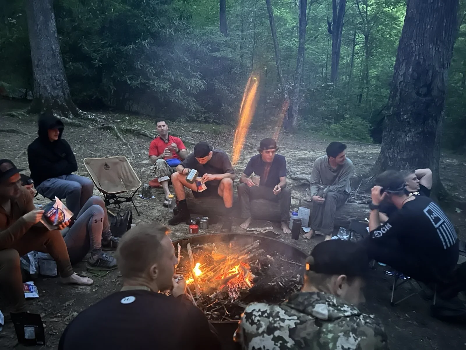 A group of people gathered around a campfire in a wooded area. They are sitting on logs and camping chairs, some holding snacks and drinks, with trees and green foliage in the background.