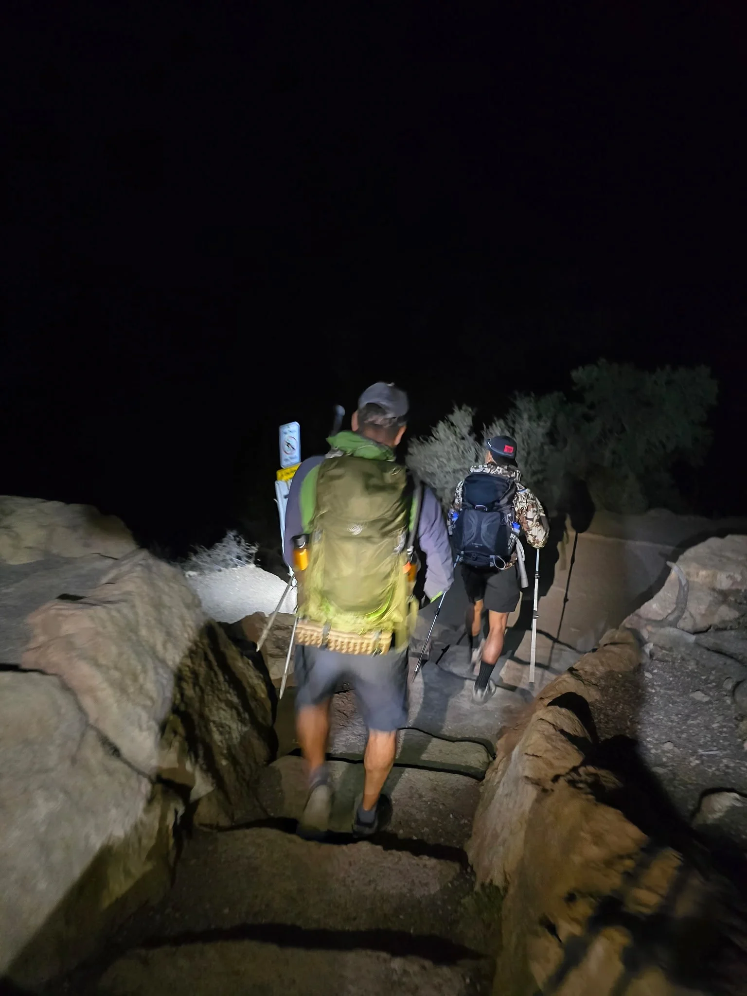 Two hikers walking down rocky stairs at night with headlamps, one wearing a green backpack and the other a black backpack.