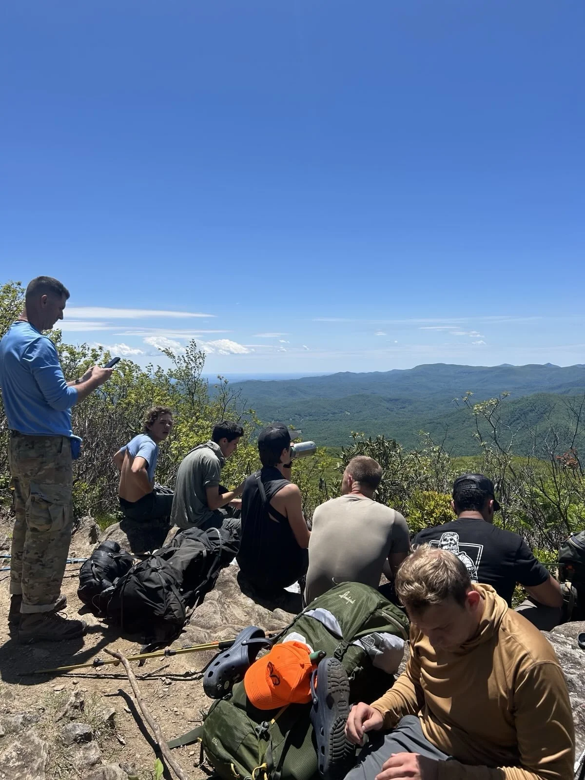 A group of hikers resting on a mountain ridge with backpacks and gear, overlooking a lush green valley and distant hills under a blue sky.