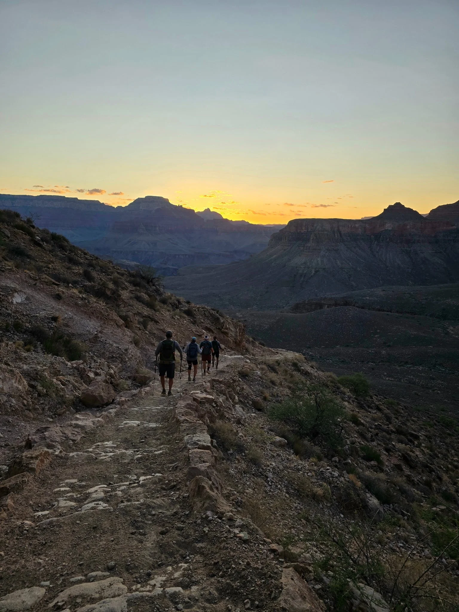 A group of hikers walking along a trail in a desert landscape at sunset, with the Grand Canyon in the background.
