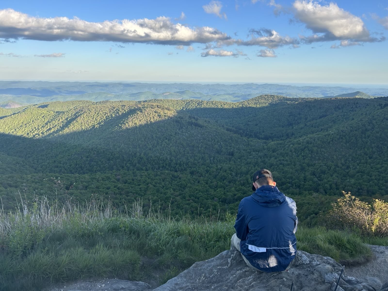 Person sitting on a rock at the edge of a mountain overlooking a vast green mountain range under a partly cloudy sky.