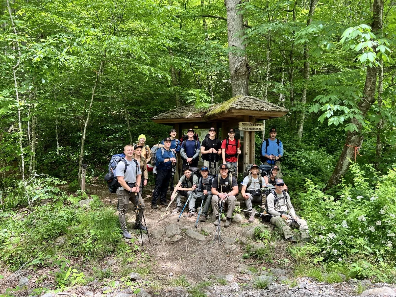 Group of hikers with backpacks and trekking poles posing in front of a small wooden shelter in a lush green forest.