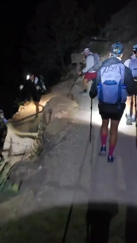 Group of hikers walking on a narrow trail at night with backpacks and walking sticks, illuminated by flashlights.