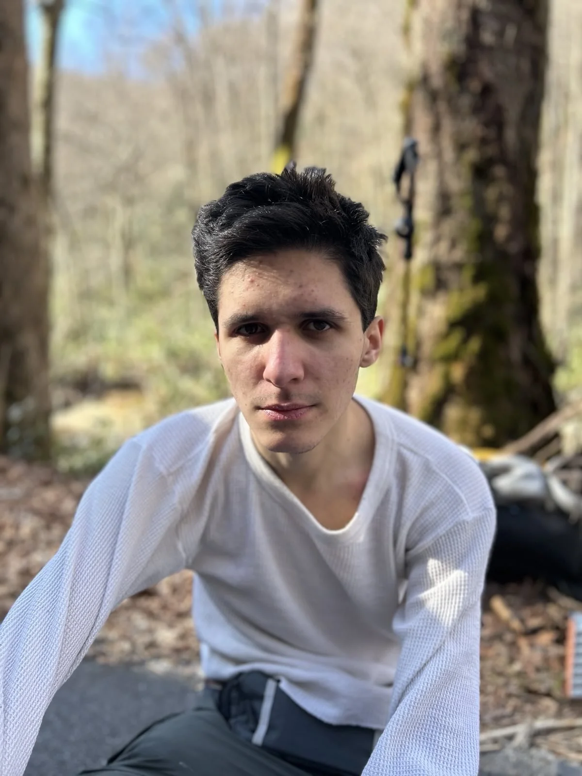 Young man in a white long sleeve shirt outdoors in a wooded area with trees and moss in the background.
