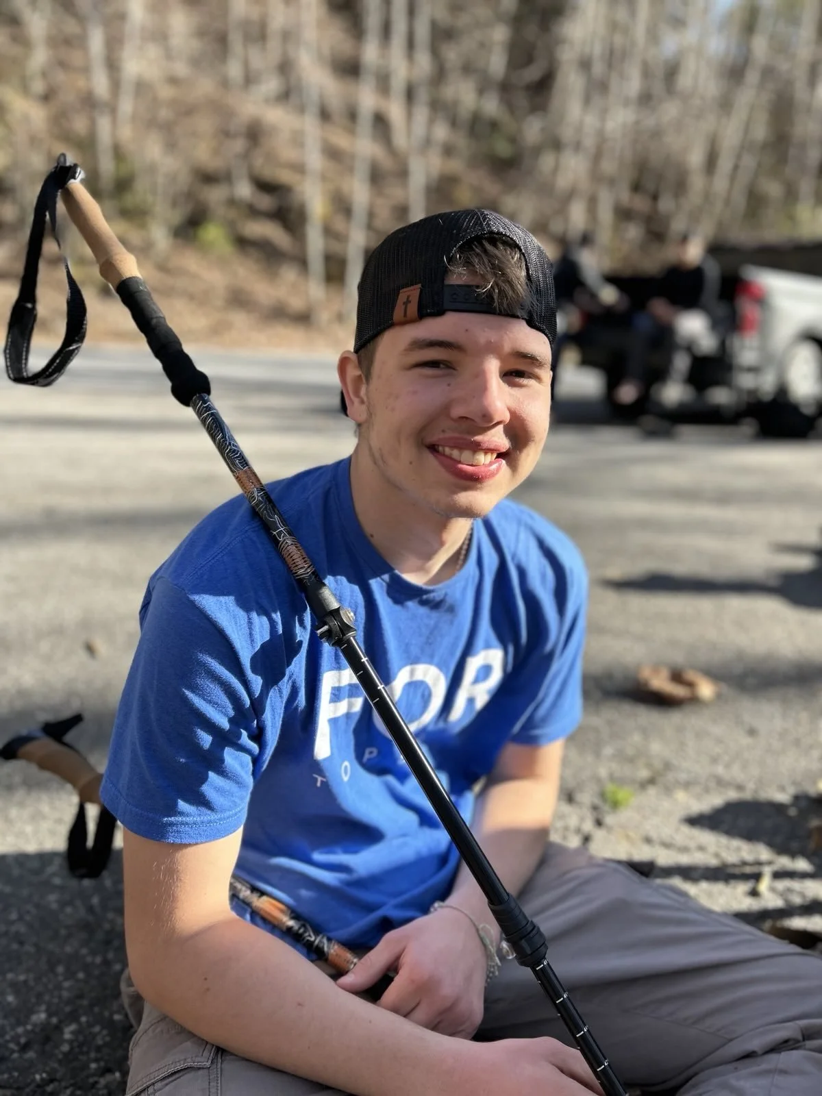 A young man sitting on the ground outdoors, smiling at the camera, wearing a blue T-shirt, a black baseball cap worn backwards, and holding a hiking stick. In the background, there are other people near a vehicle and trees.